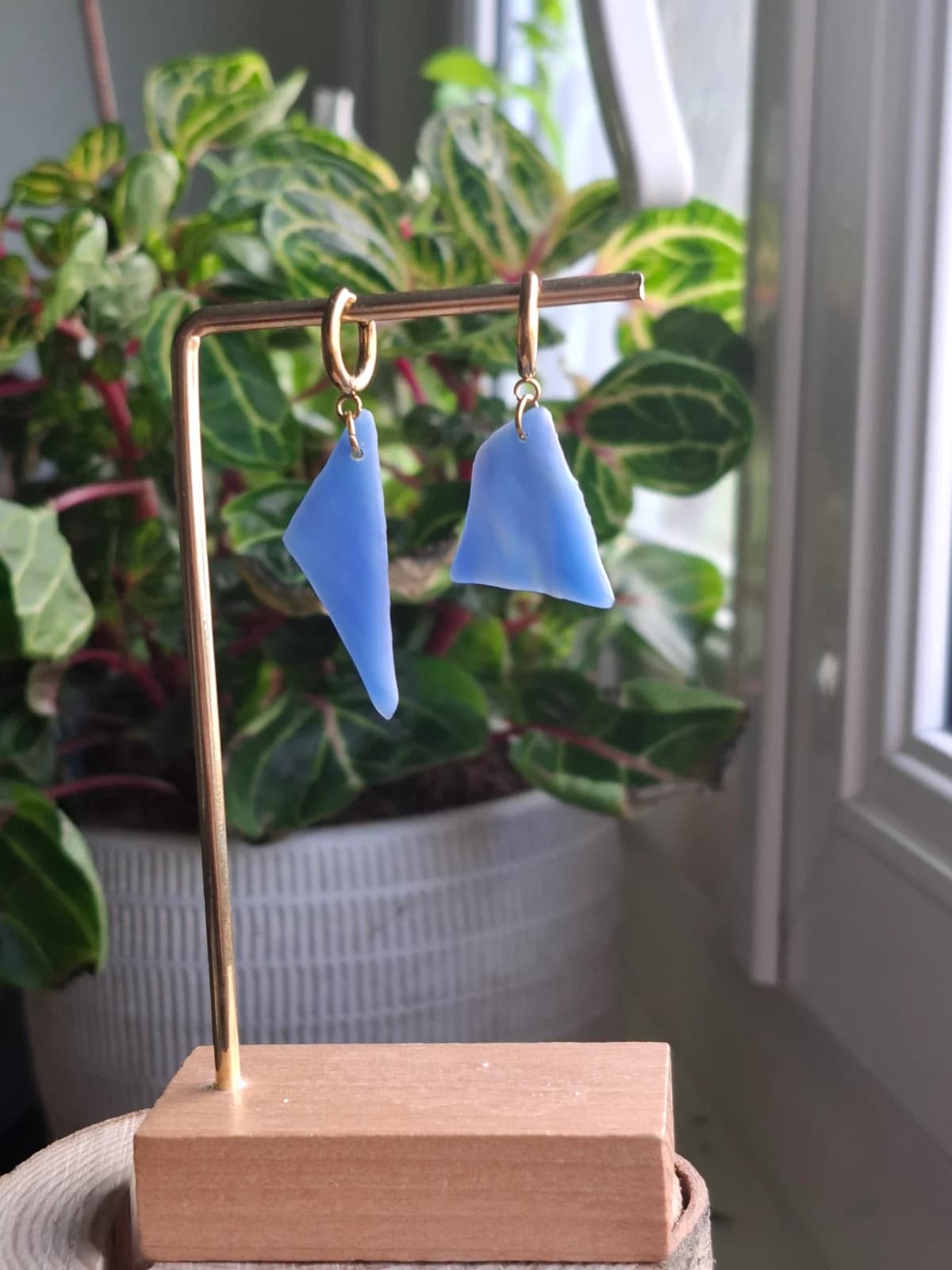 Boucles d'oreilles géométriques en verre bleu suspendues à un support doré devant une plante verte.