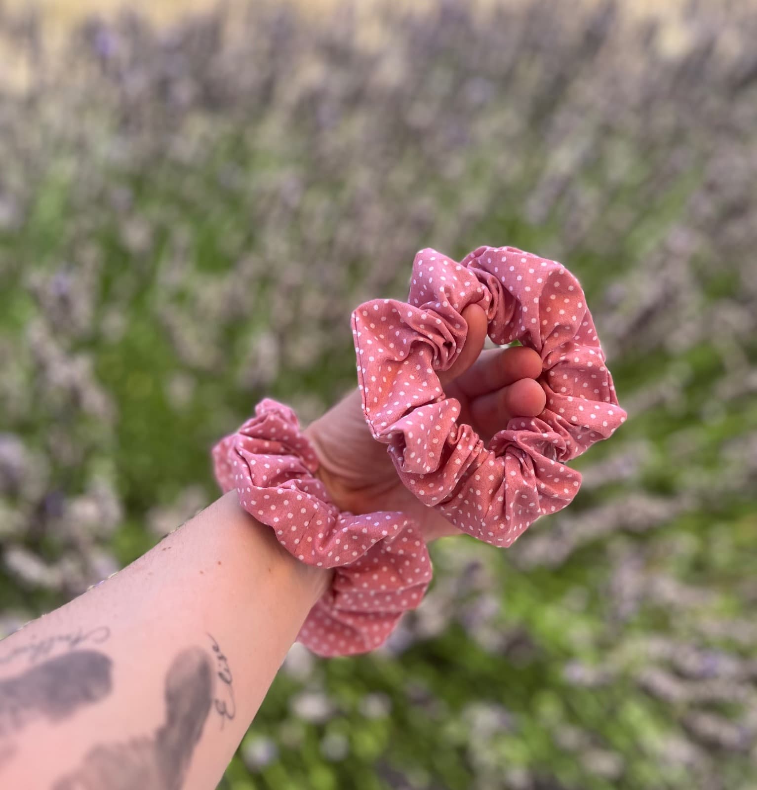 Deux chouchous en tissu rose à pois blancs tenus au poignet en extérieur devant des lavandes.